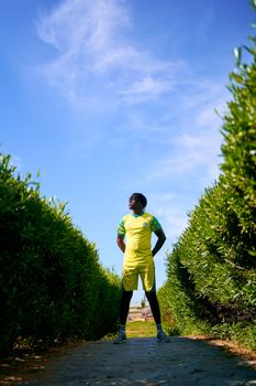 A man in a yellow soccer kit stands confidently between hedges against a clear blue sky.