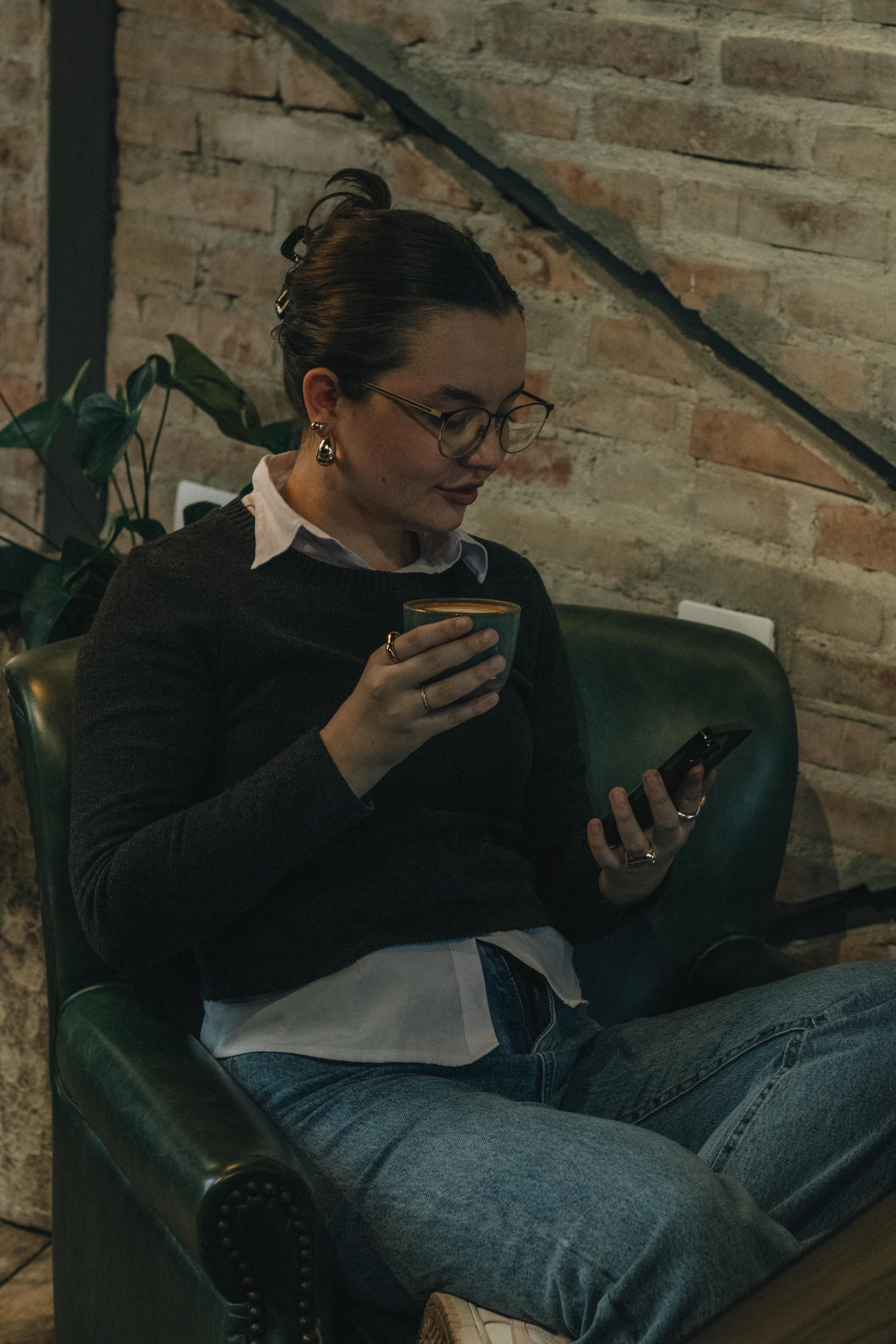 Kostnadsfria En ung kvinna sitter på ett mysigt kafé och njuter av kaffe medan hon surfar på sin smartphone. Stock foto