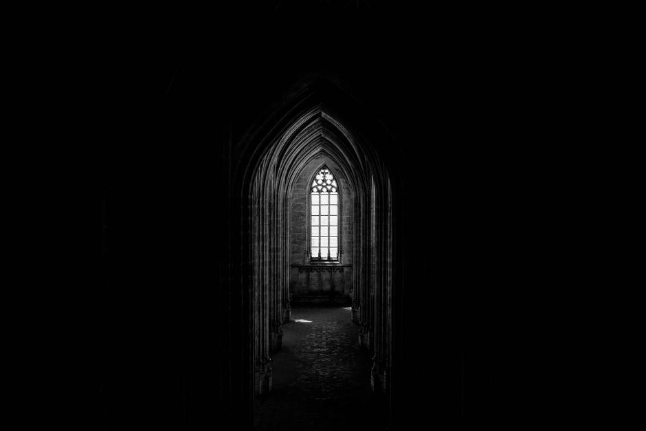 Black and white of ancient dark church with narrow corridor and arched glass window with ornament in old stone building