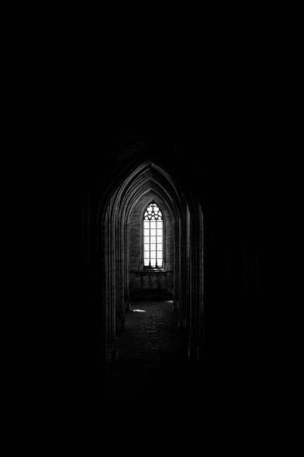 Black and white of aged stone church with small arched window in distance of dark corridor of old medieval building