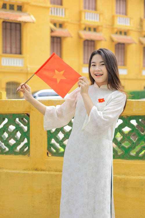 Free Young woman in traditional ao dai holding Vietnamese flag, smiling outdoors. Stock Photo