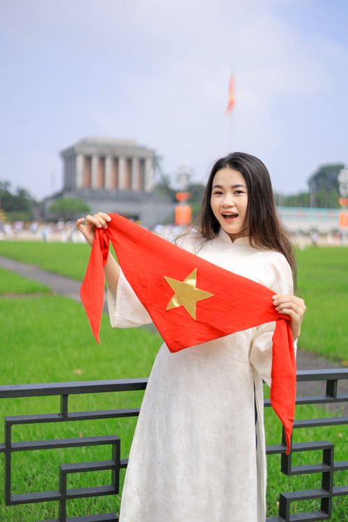 Free Vietnamese woman holding a flag in front of Ho Chi Minh Mausoleum, Hanoi, showcasing national pride. Stock Photo