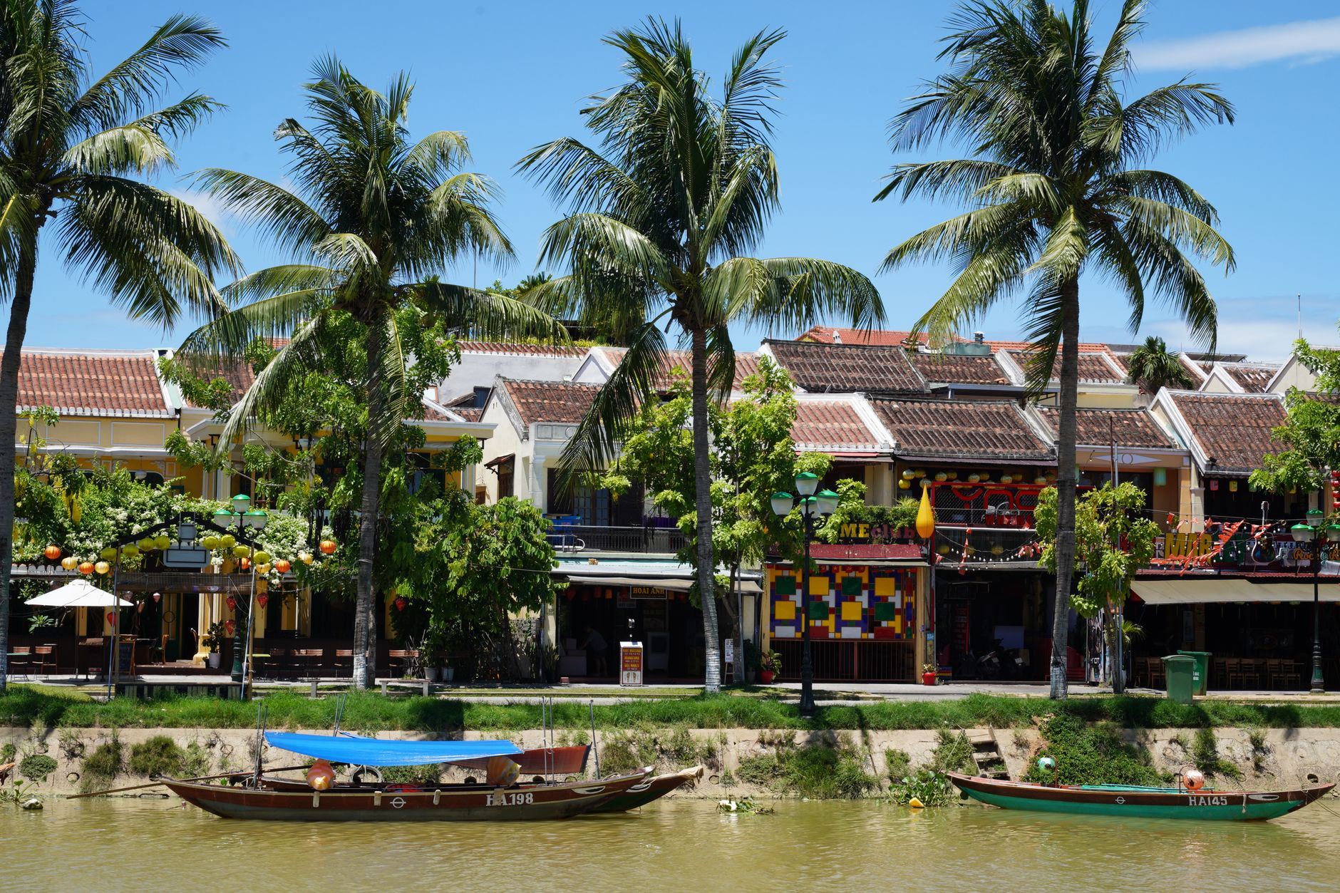 Colorful riverside town with lush palm trees and quaint boats, under a bright blue sky.
