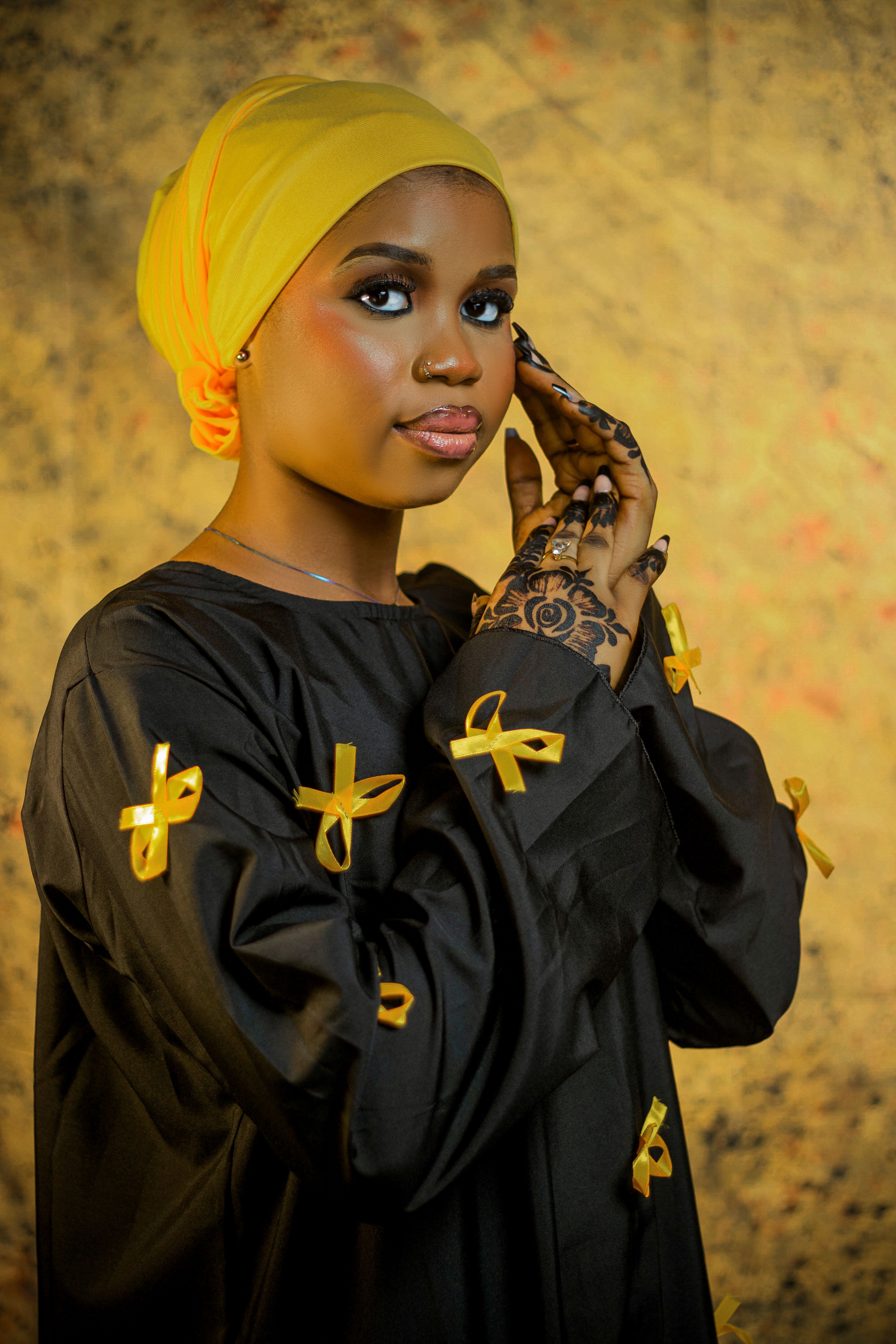 Portrait of a woman adorned with henna, wearing a yellow hijab and black dress with yellow ribbons.