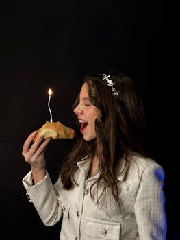A woman in a white jacket celebrates with a lit candle on a croissant, smiling against a dark background.