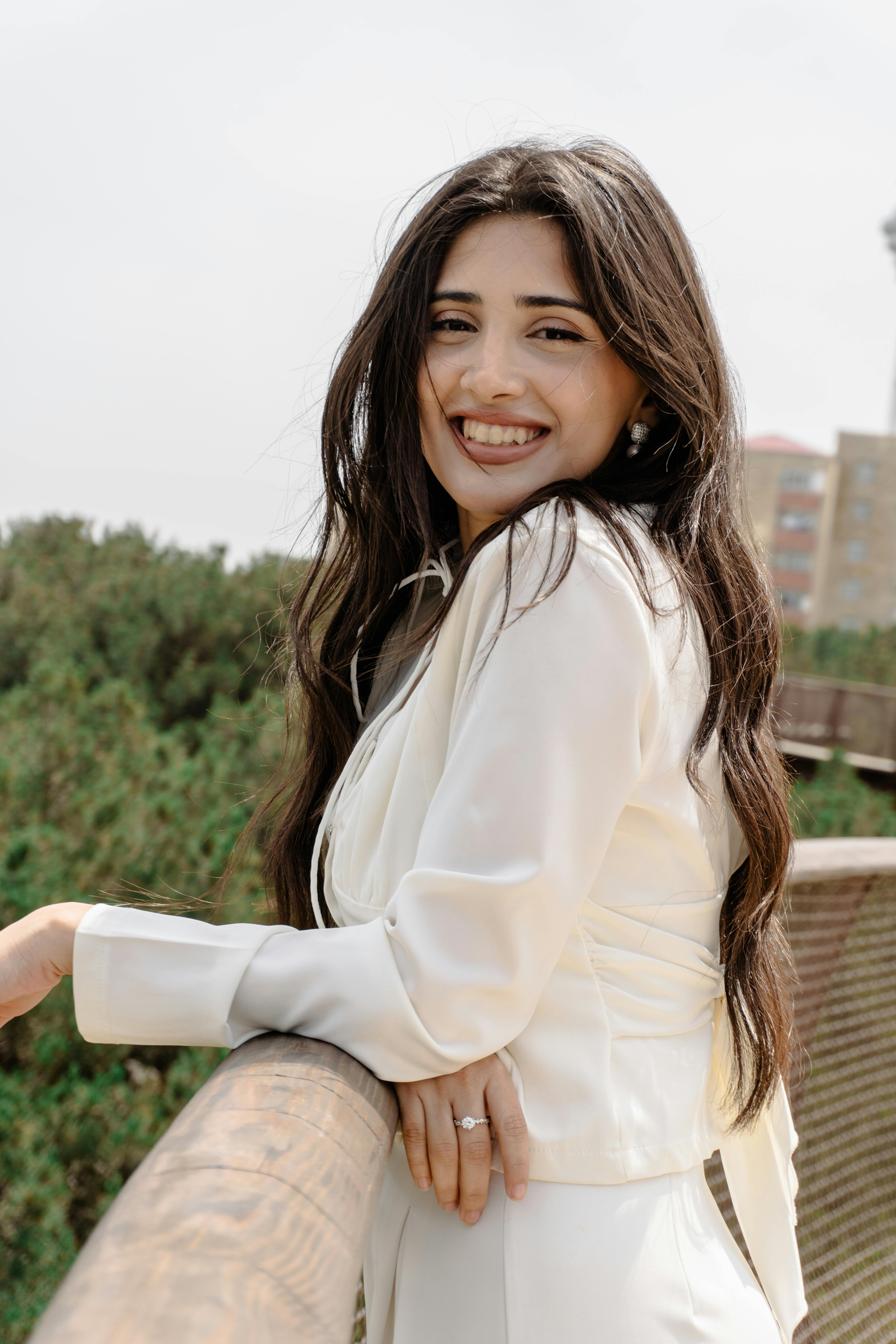 A young woman in a white outfit smiling while leaning on a wooden railing outdoors.