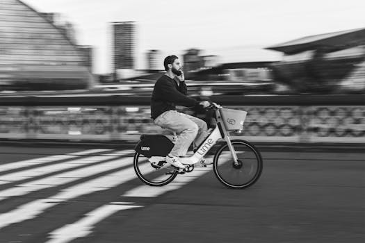 A man rides an electric bike with motion blur effect across a city bridge, in black and white.