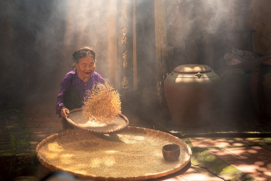 Elderly woman in Hanoi, Vietnam, sorting grains outside under sunlight.