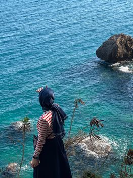A woman with a hijab gazes at the ocean from a cliff, admiring the turquoise waves.