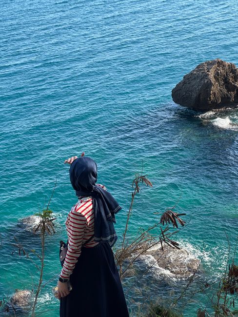 A woman with a hijab gazes at the ocean from a cliff, admiring the turquoise waves.