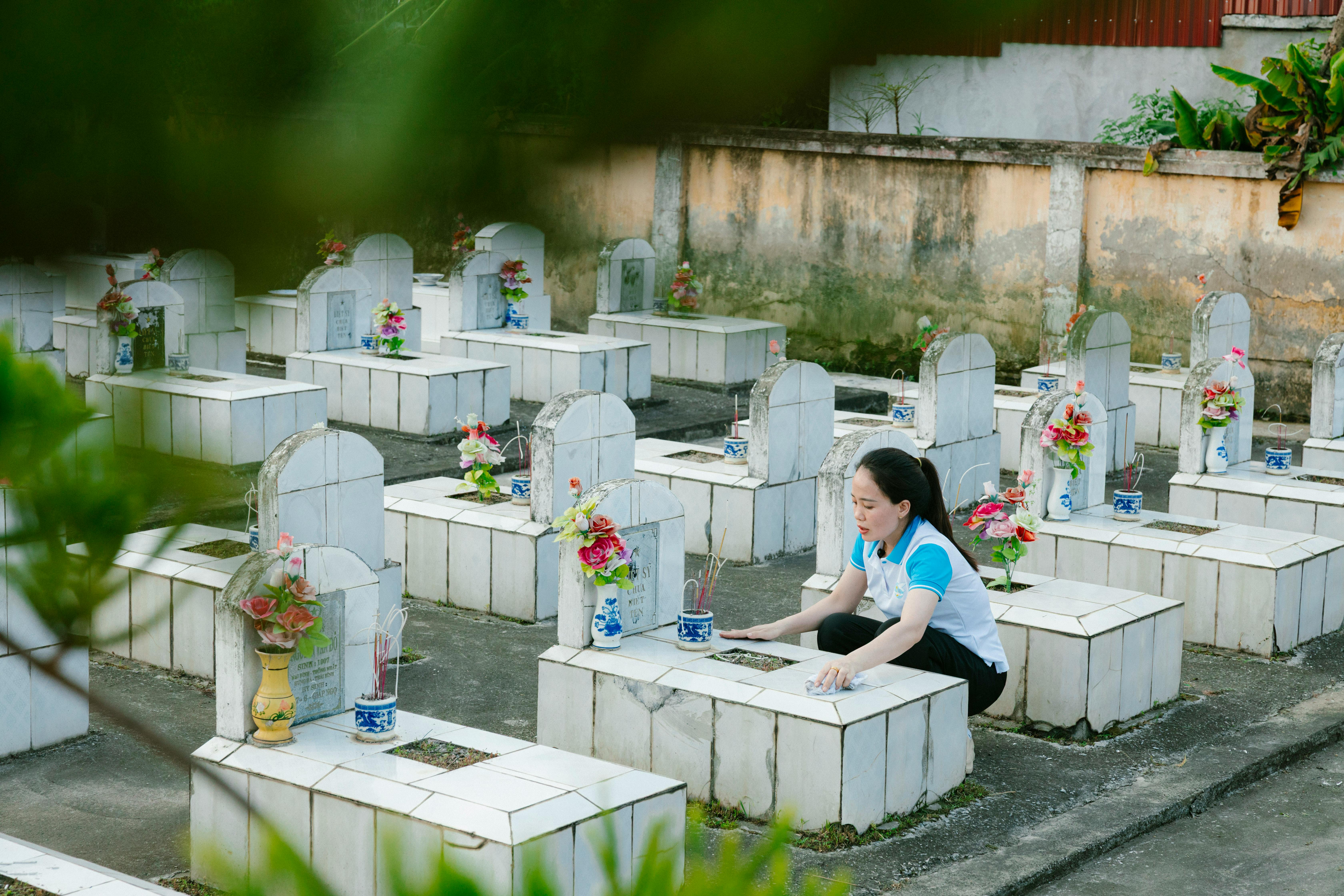 A woman tending graves with flowers and incense in a serene cemetery during the day.