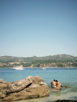 A romantic couple embraces on rocks by the sea in Pintens, Spain, under a clear summer sky.