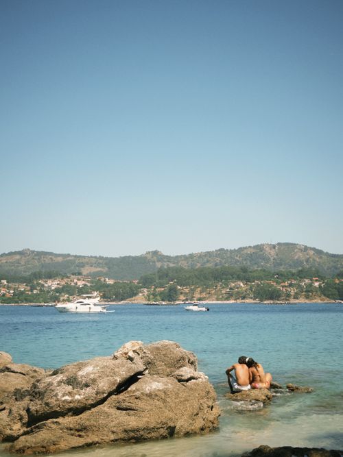 Free A romantic couple embraces on rocks by the sea in Pintens, Spain, under a clear summer sky. Stock Photo