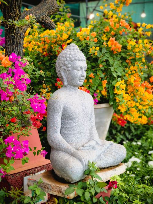 Free Peaceful Buddha statue surrounded by lush, colorful flowers in a tranquil garden setting. Stock Photo
