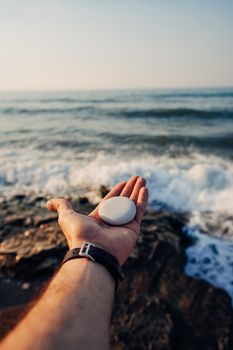 A hand holding a smooth stone by the seaside with waves crashing in the background.