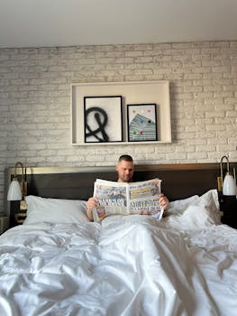 Caucasian man in a hotel room reading a newspaper while relaxed in bed with minimalistic decor.