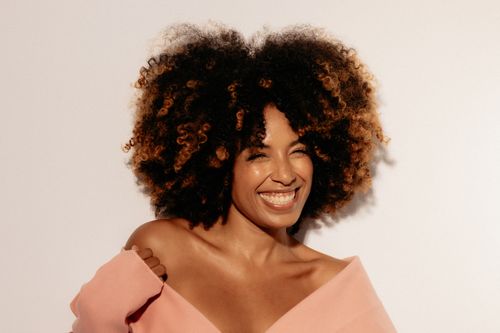 Free Cheerful woman with curly afro hair in pink attire, smiling brightly against a neutral background. Stock Photo