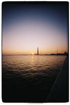 Sunset over calm waters with silhouette of a tall landmark in the background.