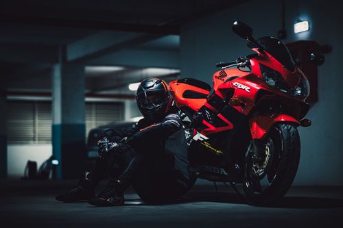 Free Motorcyclist in full gear sits in a dimly lit garage next to a vibrant red sport bike, creating a dramatic atmosphere. Stock Photo