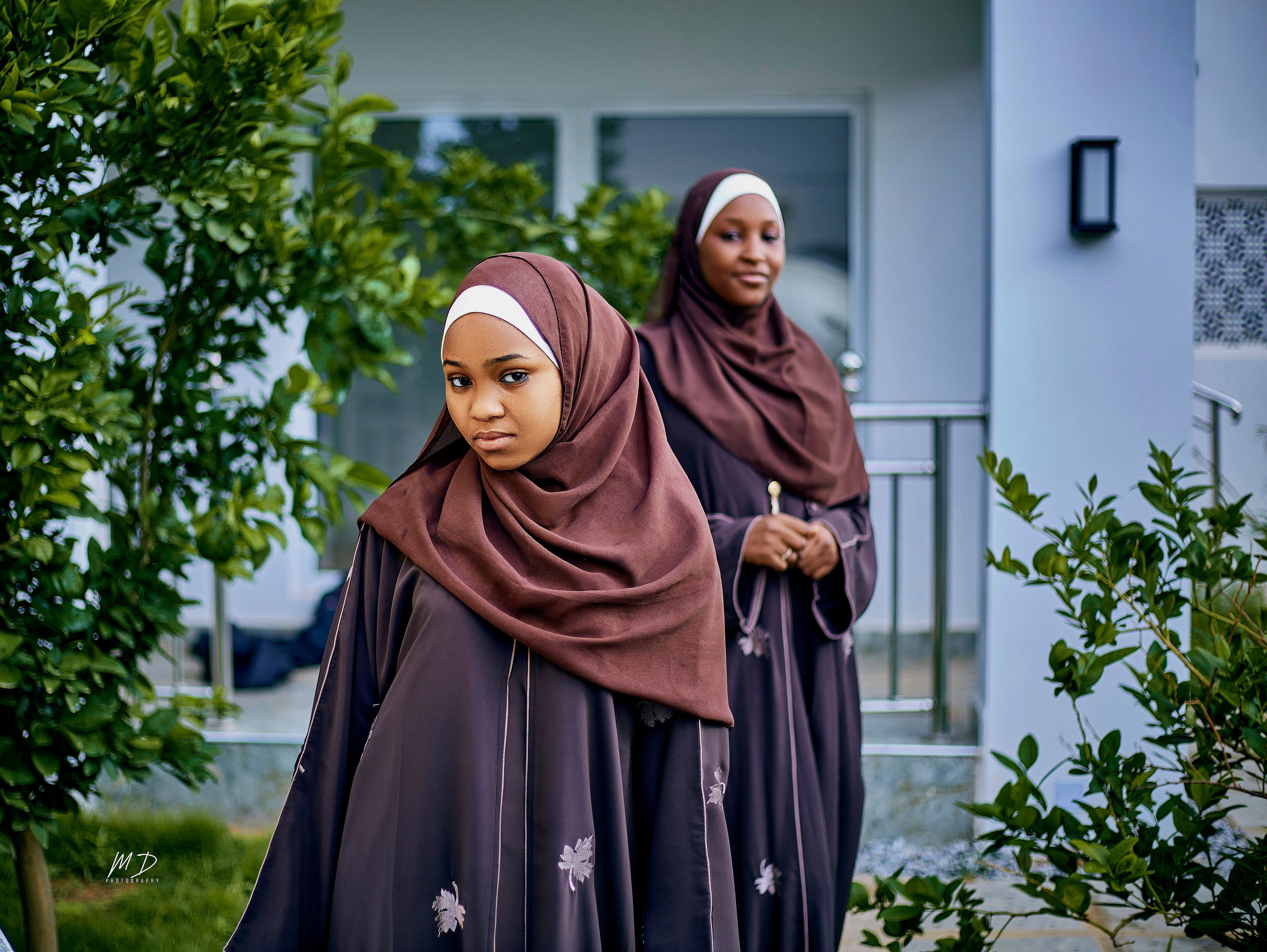 Two women in brown hijabs standing elegantly outdoors near green foliage.
