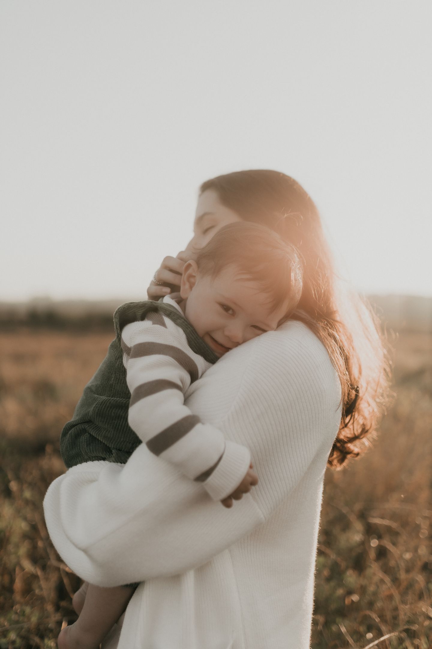 A mother holding her young child together in warm, sunlit meadow light.