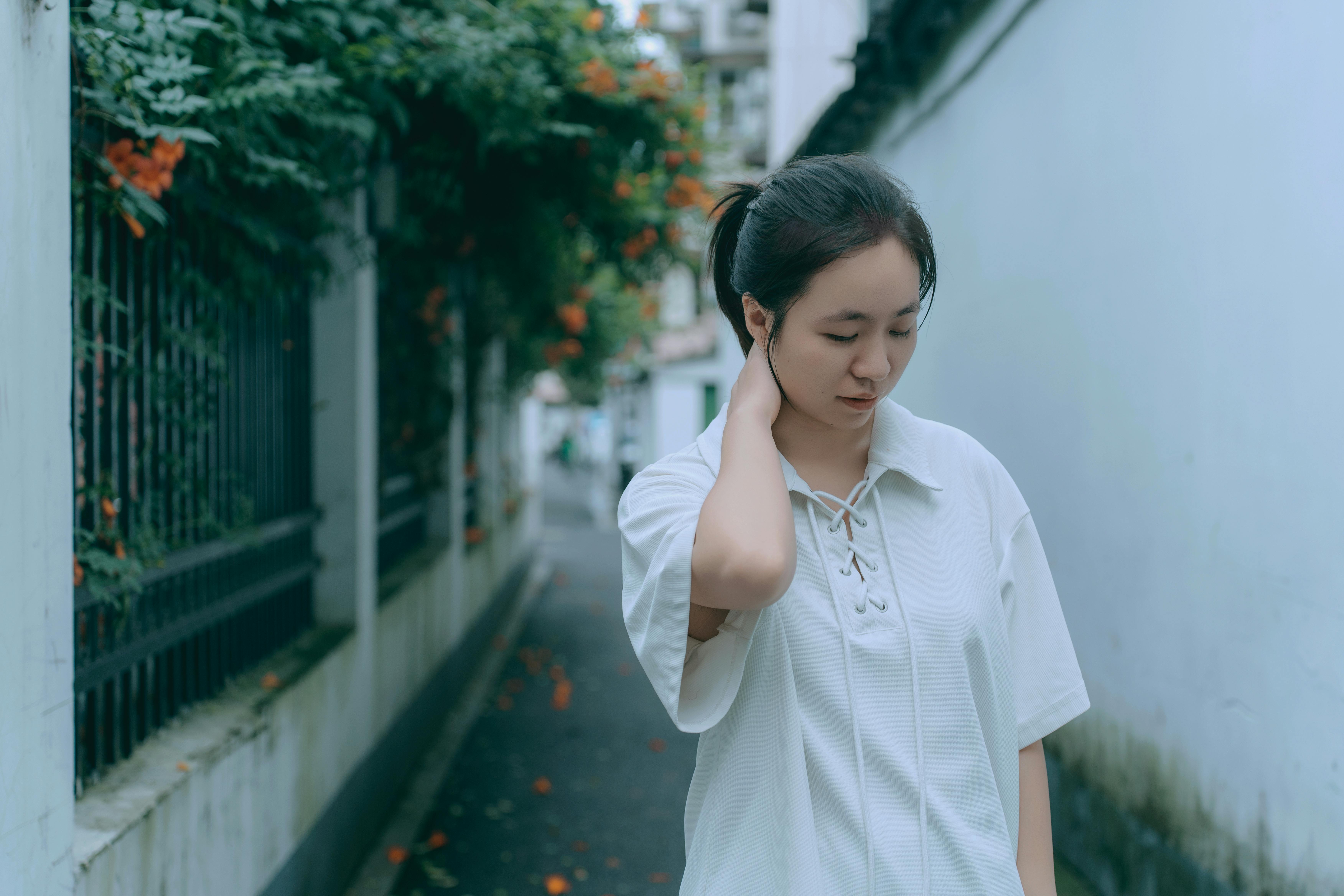 A woman in a white shirt ponders quietly in a narrow alley lined with greenery and flowers.