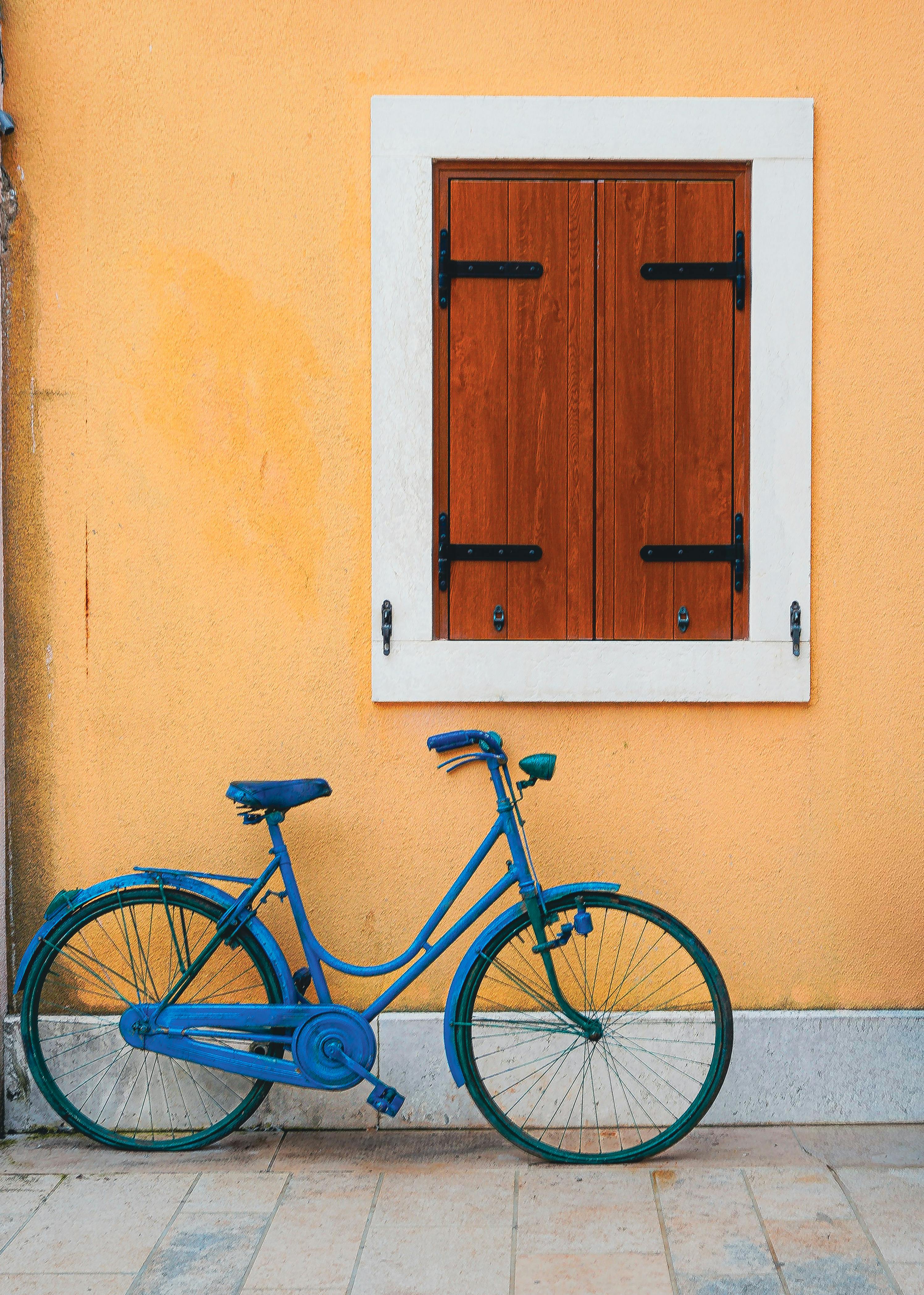 A vibrant blue bicycle leans against an orange wall under a rustic wooden window. Perfect for travel and lifestyle themes.