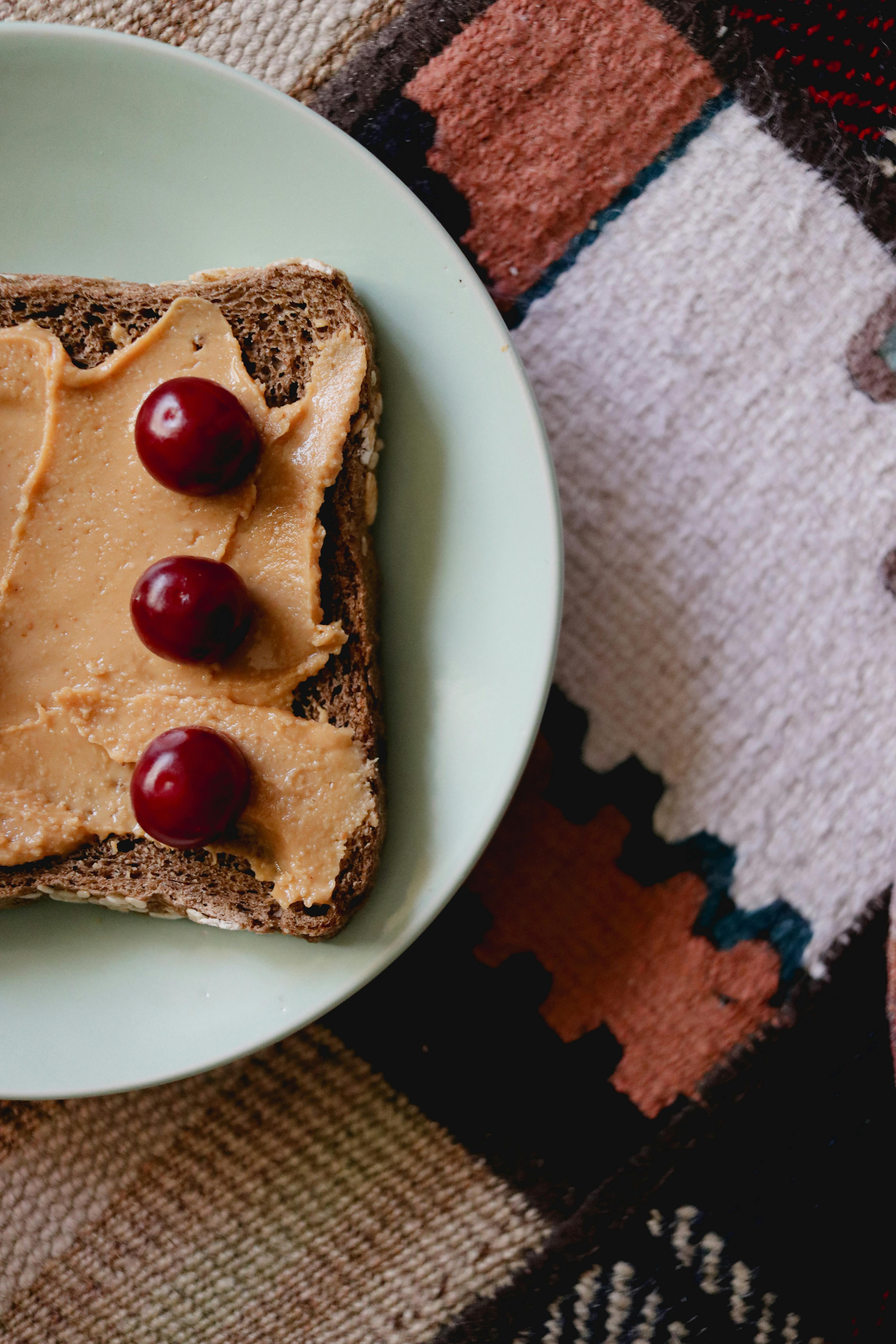Close-up of wholegrain toast with peanut butter and cherries on patterned mat.