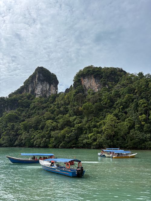 Free Long boats float on turquoise waters near lush cliffs under a cloudy sky. Stock Photo