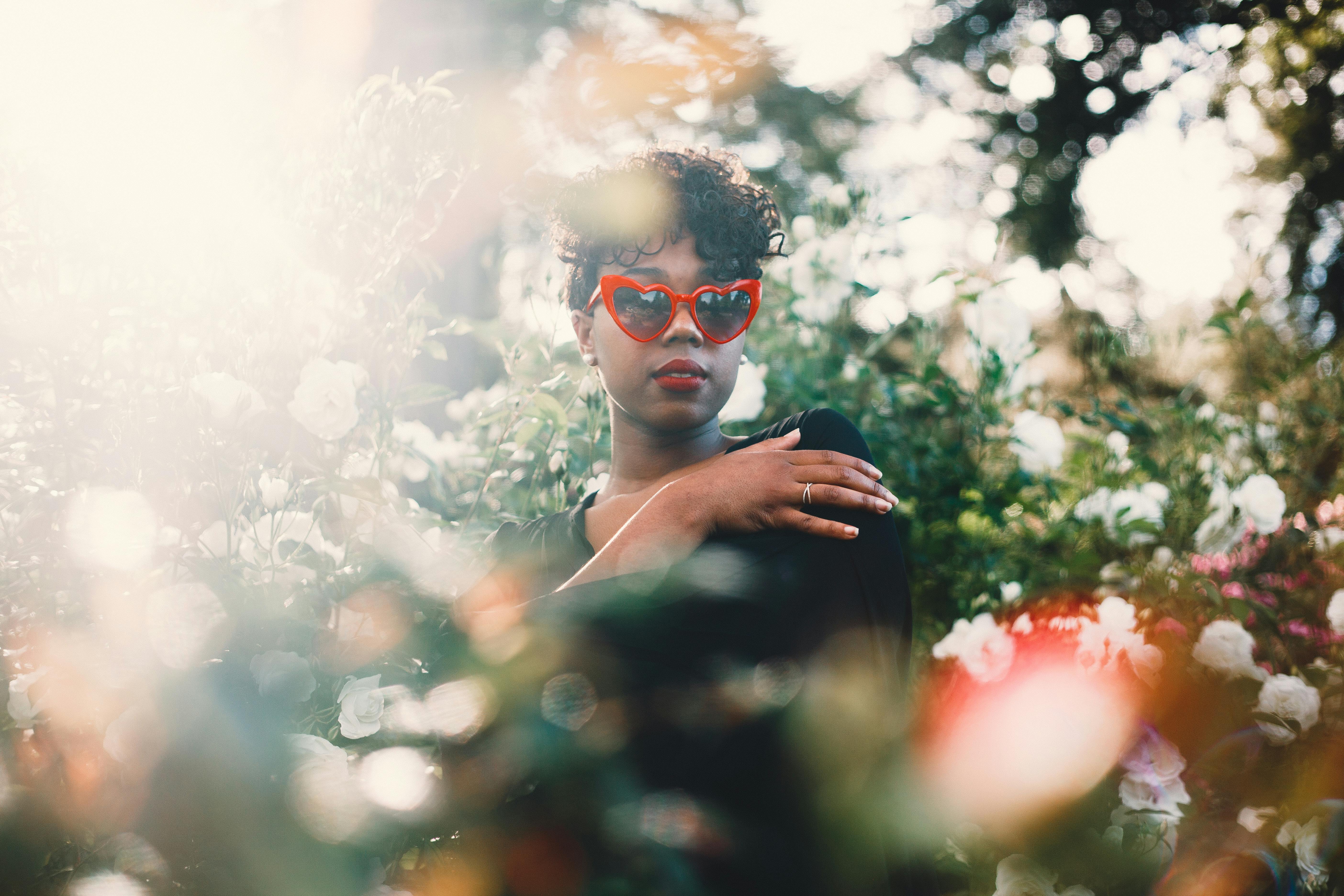 一位戴着红色心形太阳镜的女性肖像，在阳光明媚的花园中被盛开的花朵环绕