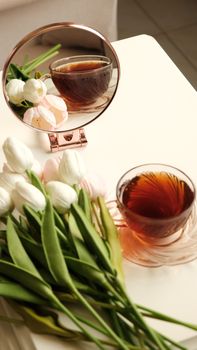 A serene setup of tea and tulips reflected in a mirror on a white table.
