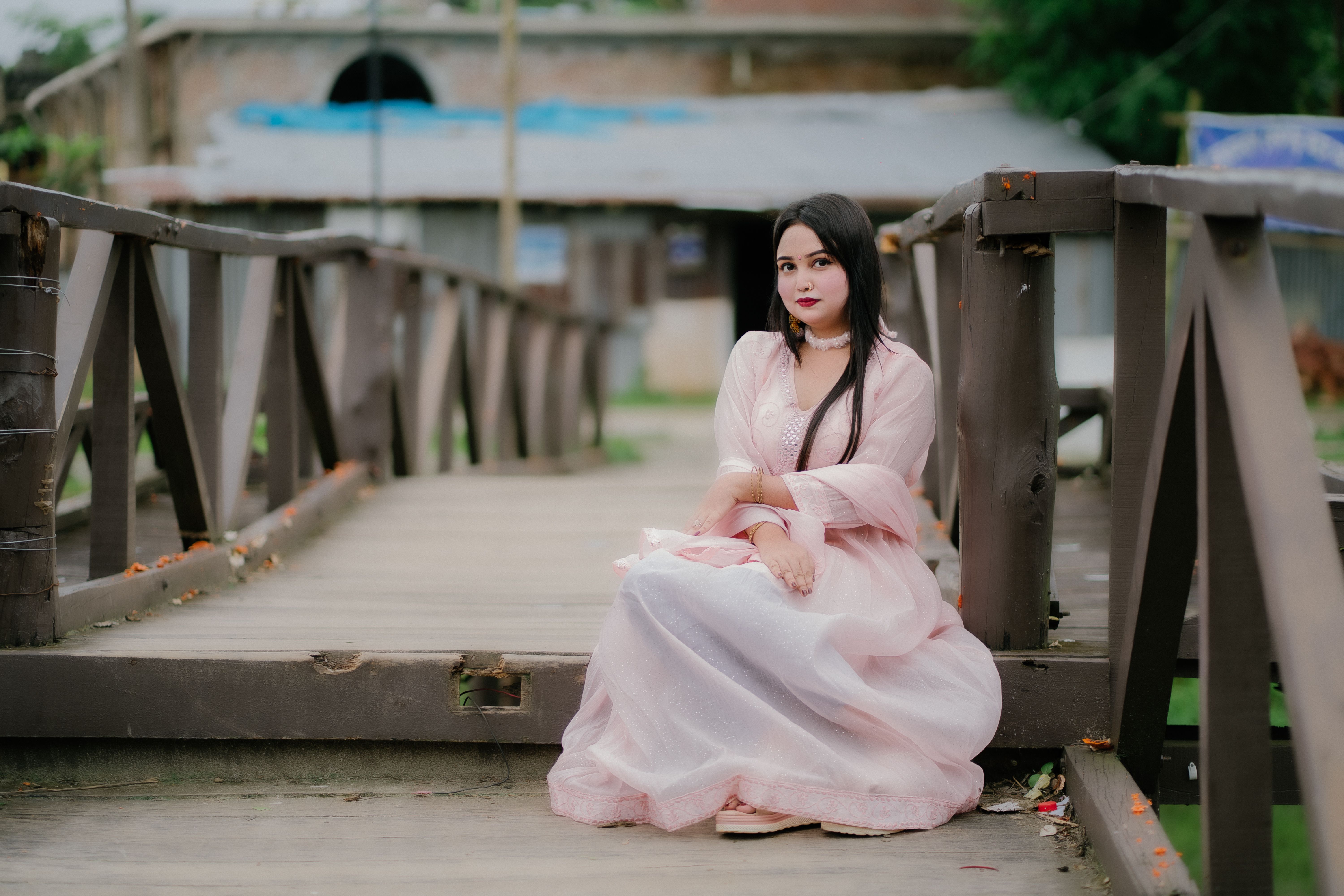 Young woman in pink traditional dress sitting on a rustic wooden bridge outdoors.
