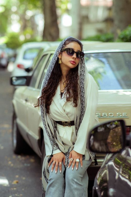 Free A fashionable woman poses confidently next to a vintage car on a Tehran street. Stock Photo