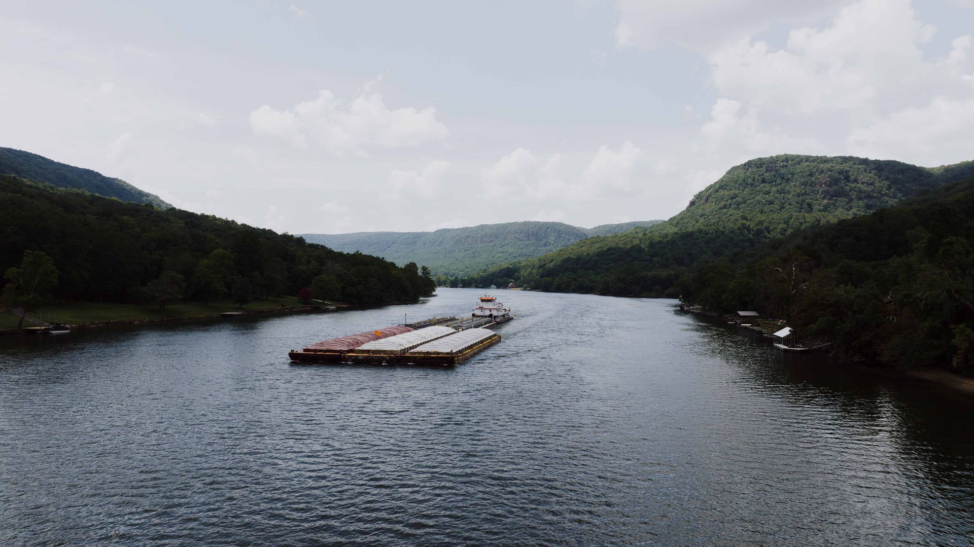 A serene aerial shot of a barge navigating the Tennessee River surrounded by lush hills and summer skies.