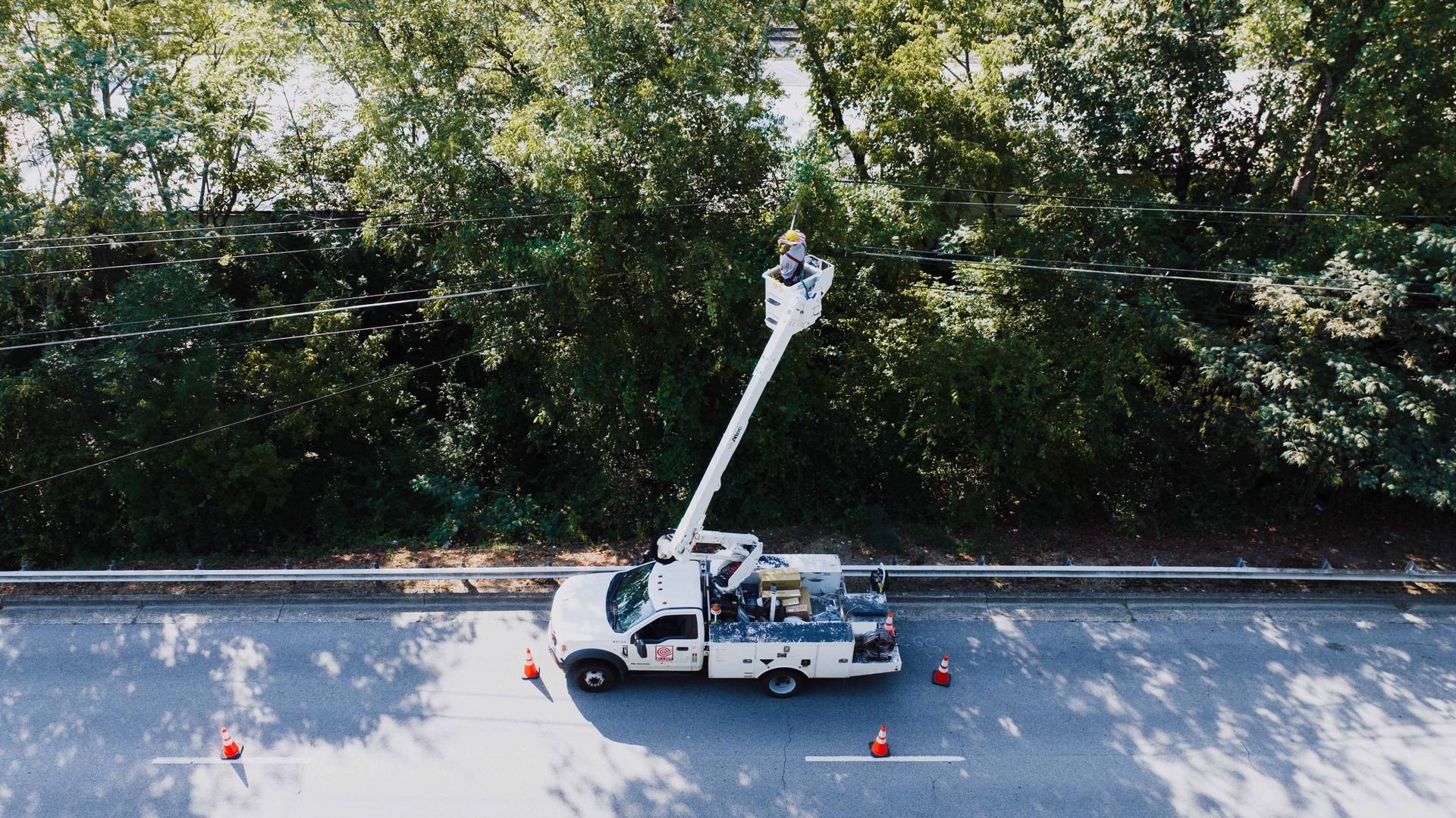 Aerial view of a utility worker on a bucket truck fixing power lines by a road.