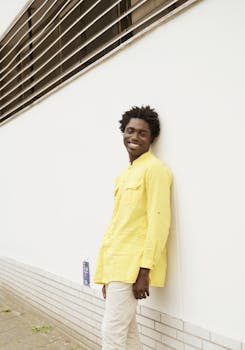 A smiling man in a yellow shirt leans against a modern Berlin building, showcasing minimal style.