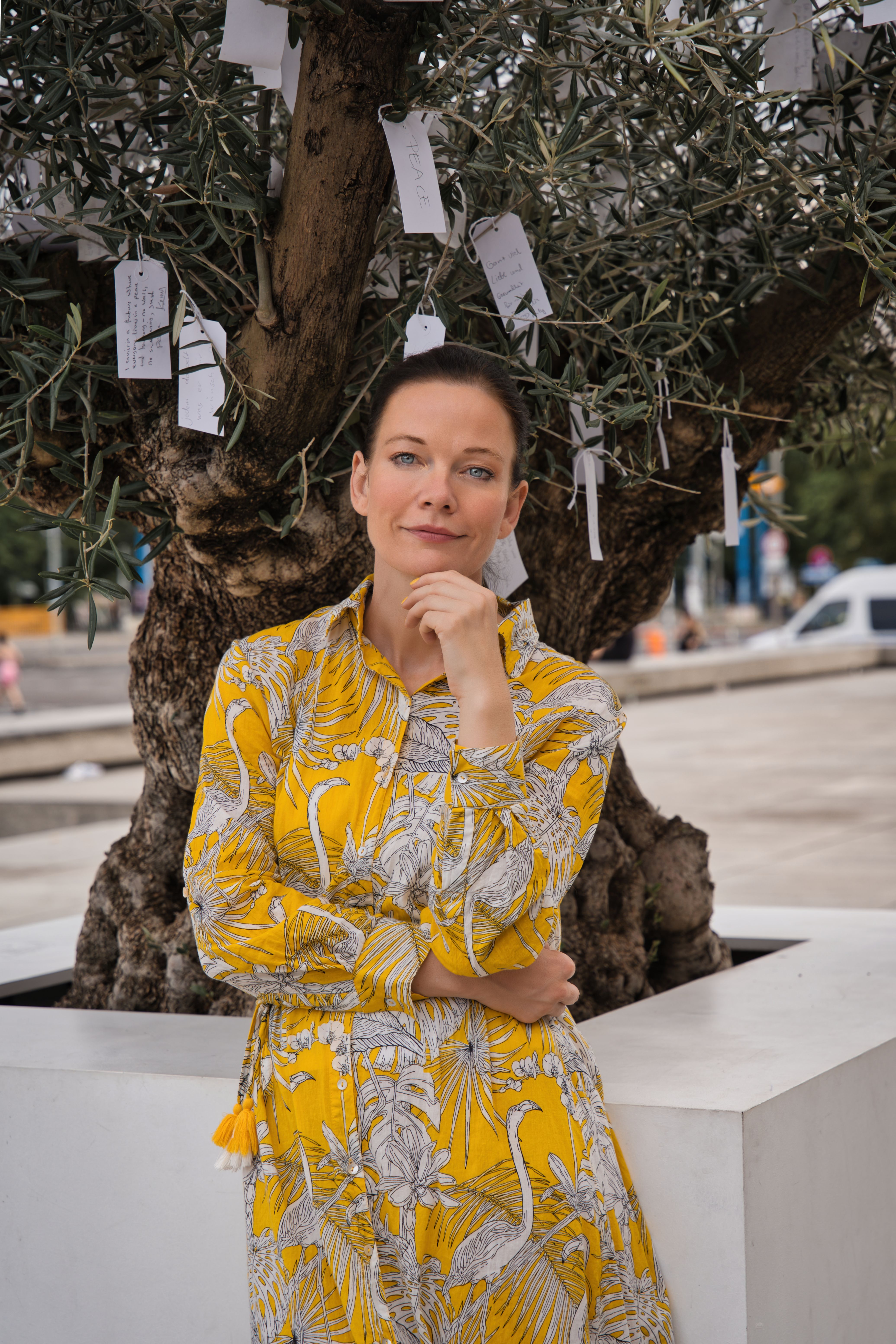 Portrait of a woman in a yellow dress standing by a wishing tree adorned with notes in Berlin.