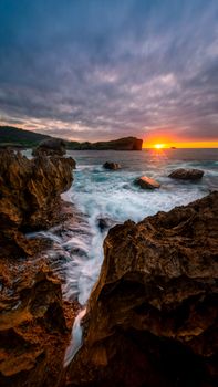 Vibrant sunset over rocky shoreline with crashing waves and dramatic clouds in the sky.