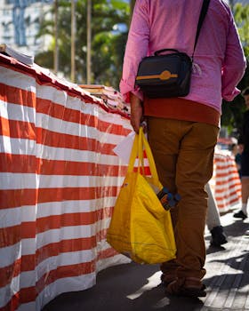 A shopper carrying bags at an outdoor market with striped tents, under daylight.
