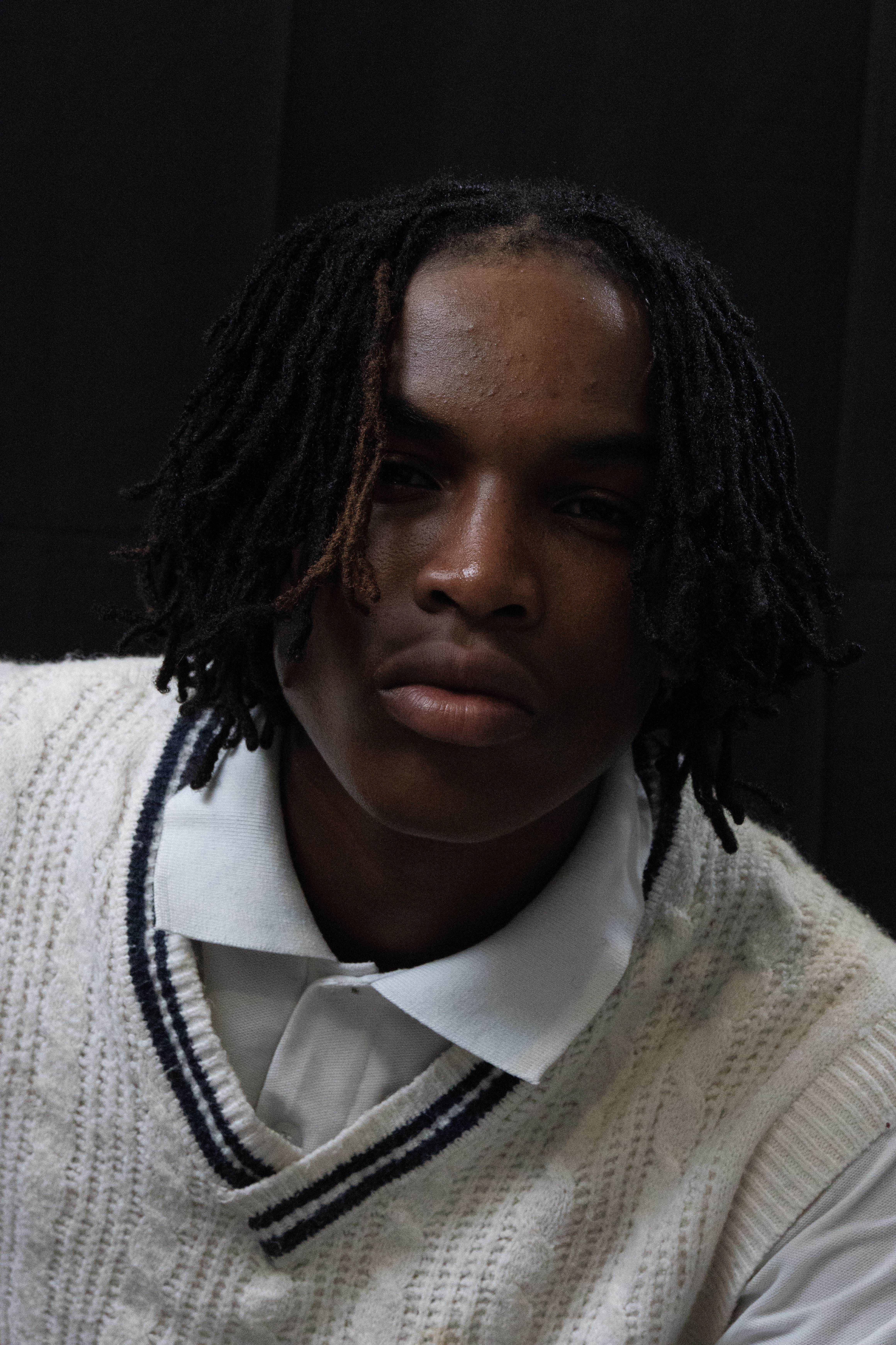 Close-up portrait of a young man in a sweater with dreadlocks, posing indoors.