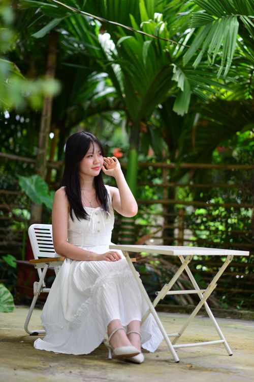 Free Asian woman in a white dress sitting at a table in a lush tropical garden, enjoying the serene outdoor setting. Stock Photo