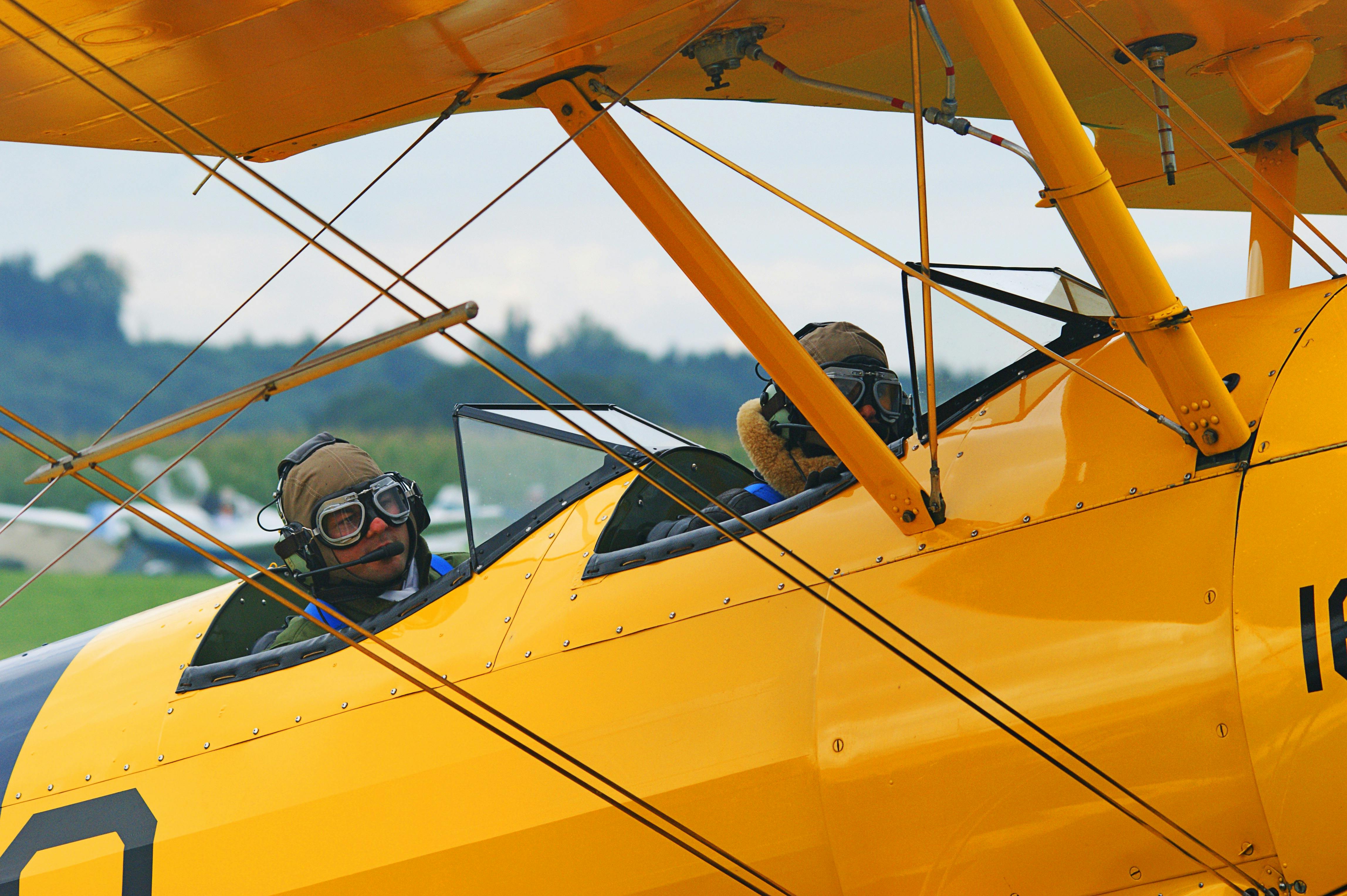 Two pilots in vintage yellow biplane with open cockpit, flying over countryside.