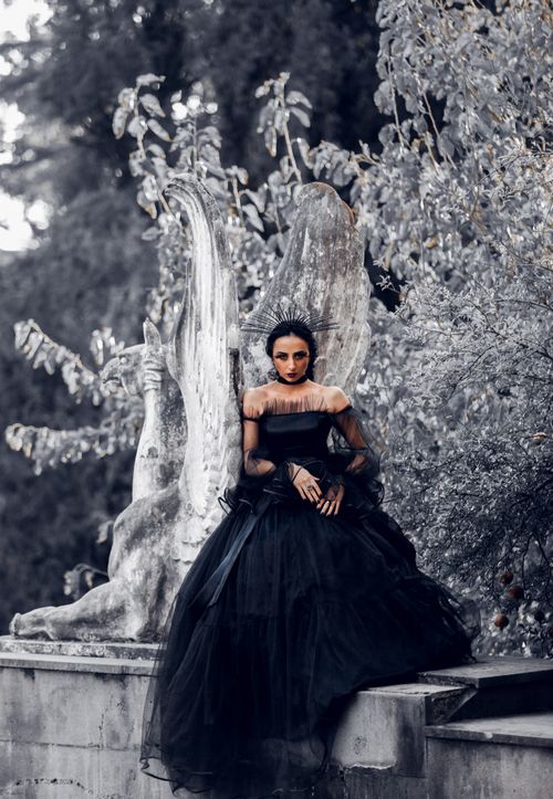 Free A captivating portrait of a woman in a black gown sitting beside a marble statue outdoors. Stock Photo