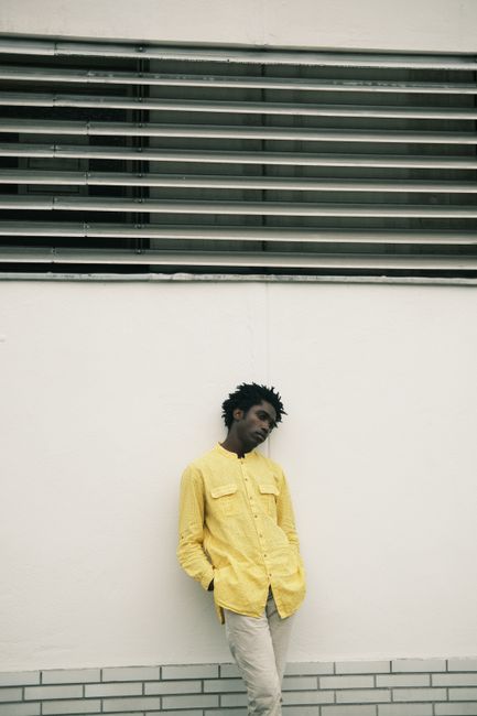 Contemplative man in yellow shirt leaning against a wall outdoors, urban setting.