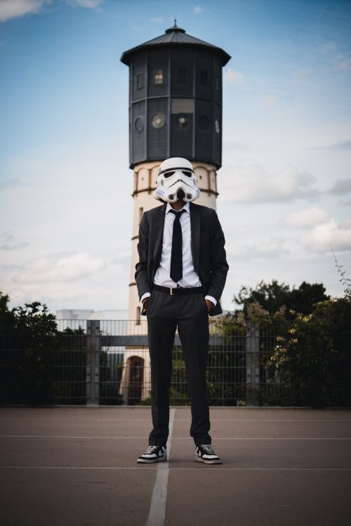 Free A man in a suit wearing a Stormtrooper helmet stands outside near a water tower. Stock Photo