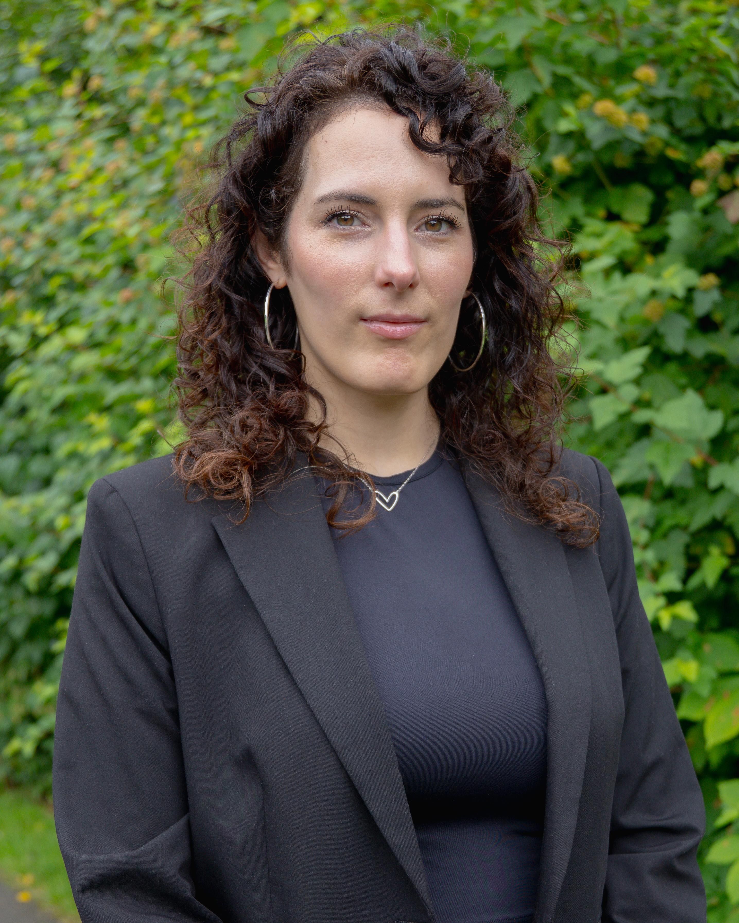 Confident woman with curly hair posing in a black suit outdoors.