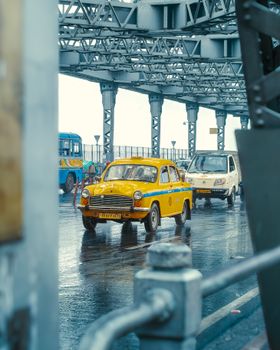 A classic yellow taxi navigates rainy weather on the historic Howrah Bridge in Kolkata, India.
