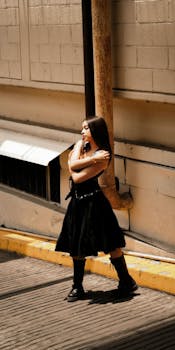 Stylish woman posing in a sunlit urban alleyway, wearing a black dress and boots.