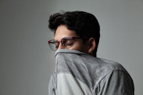 Free Young man in glasses covers mouth with sweater, lost in thought. Stock Photo