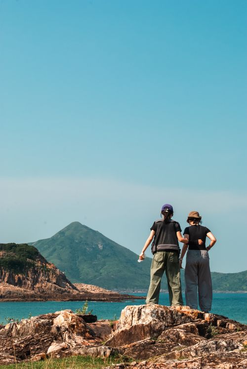 Free Two hikers stand on rocky terrain overlooking the sea towards a distant mountain under a clear blue sky. Stock Photo
